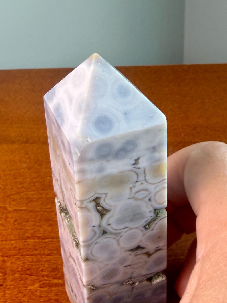 May include: A polished stone tower with a pointed top, featuring a natural pattern of circular, orb-like inclusions in shades of pale blue, white, and soft beige, with small crystalline crevices visible along the vertical sides of the mineral.