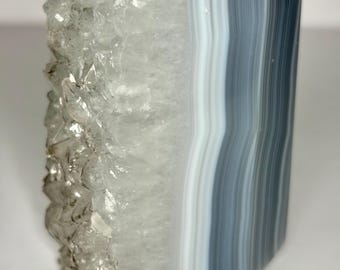 May include: A polished agate crystal tower featuring a distinct split design. One side displays a rough, translucent, and jagged crystalline texture in clear and white tones, while the other side shows smooth, parallel bands of gray, white, and charcoal.