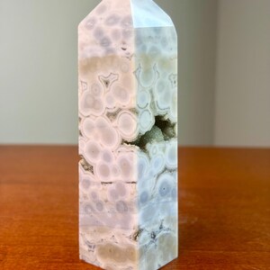 May include: A polished crystal tower featuring a light gray and white orbicular pattern with circular inclusions throughout the stone. A small, jagged horizontal opening reveals a cluster of tiny, sparkling crystals embedded within the center.