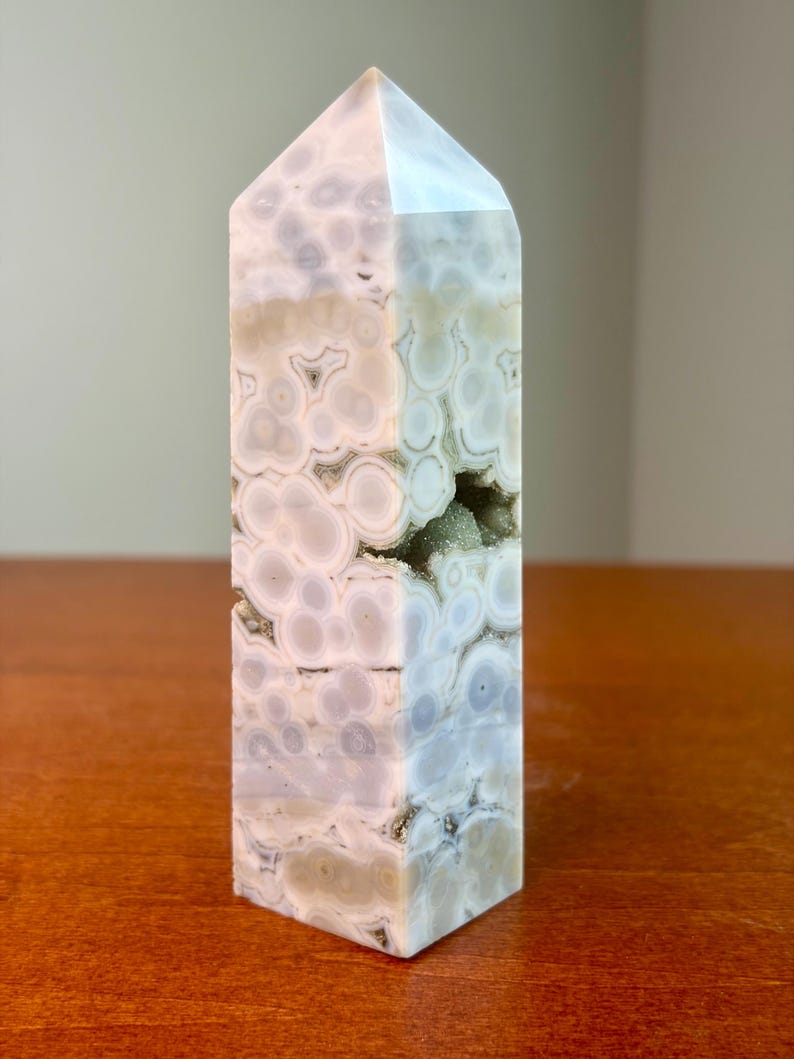 May include: A polished crystal tower featuring a light gray and white orbicular pattern with circular inclusions throughout the stone. A small, jagged horizontal opening reveals a cluster of tiny, sparkling crystals embedded within the center.