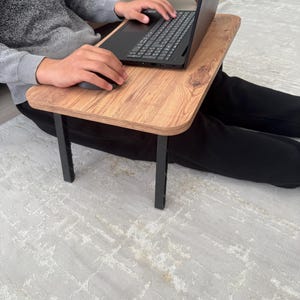 May include: A wooden laptop stand with black metal legs. The stand is being used by a person sitting on the floor.