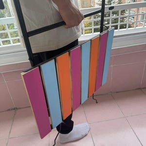 May include: A black metal folding table with a colorful striped wooden top. The table is pink, orange, blue, and light pink. The table is being held by a person.