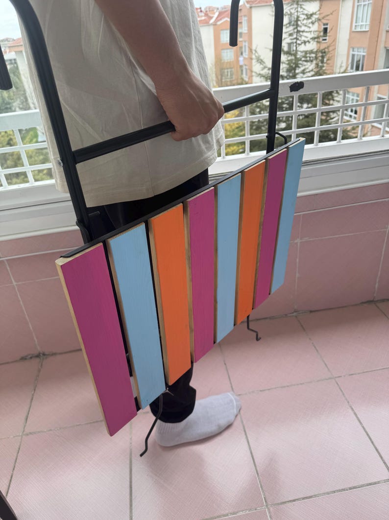 May include: A black metal folding table with a colorful striped wooden top. The table is pink, orange, blue, and light pink. The table is being held by a person.