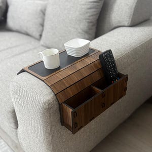 Puede incluir: Una bandeja de madera para reposabrazos con dos compartimentos para guardar objetos como un mando a distancia, una taza o un cuenco. La bandeja está unida al reposabrazos de un sofá gris.