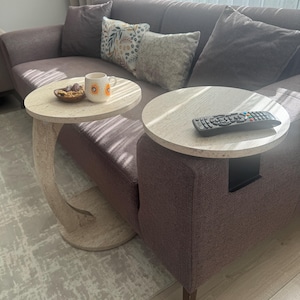 May include: Two small, round side tables with light-coloured tops are positioned next to a mauve sofa. One table holds a mug with a sunflower design and a small bowl. The other table has a remote control on top.