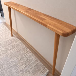 Op de afbeelding: Een lange, smalle consoletafel van lichtgekleurd hout met een glad, rechthoekig blad met afgeronde hoeken. De tafel wordt ondersteund door twee slanke, taps toelopende cilindrische poten aan elk uiteinde van de structuur.