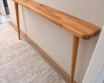Peut inclure: Une console longue et étroite en bois clair avec un plateau rectangulaire lisse aux bords arrondis. La table est soutenue par deux pieds cylindriques fins et effilés, positionnés à chaque extrémité de la structure.