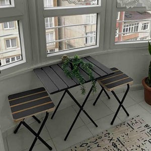 May include: A black folding table and two matching chairs with black metal frames and wooden tops with black stripes. The table and chairs are set up in a room with a window and a rug.