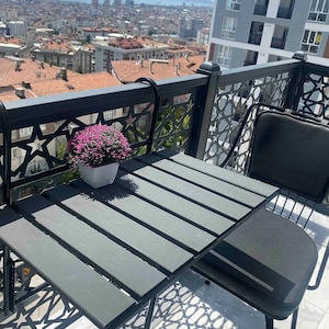 May include: A small black metal table with a grey wooden top and a black metal chair with a grey cushion. The table and chair are on a balcony with a view of a city skyline.