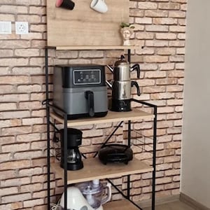 Puede incluir: Una estantería de cocina de metal negro con tres estantes de madera. El estante superior tiene un pequeño tablero trasero de madera con dos tazas colgadas de ganchos. El segundo estante tiene una tetera eléctrica plateada, una freidora de aire gris y una cafetera negra. El tercer estante tiene una plancha eléctrica negra y un procesador de alimentos blanco. La estantería está frente a una pared de ladrillo.