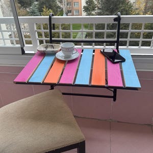 Op de afbeelding: Een kleurrijke opklaptafel met een zwart metalen frame en een roze, oranje en blauwe gestreepte bovenkant. De tafel is bevestigd aan een wit hek en heeft een klein wit kopje en schoteltje erop. Een zwarte mobiele telefoon ligt ook op de tafel.
