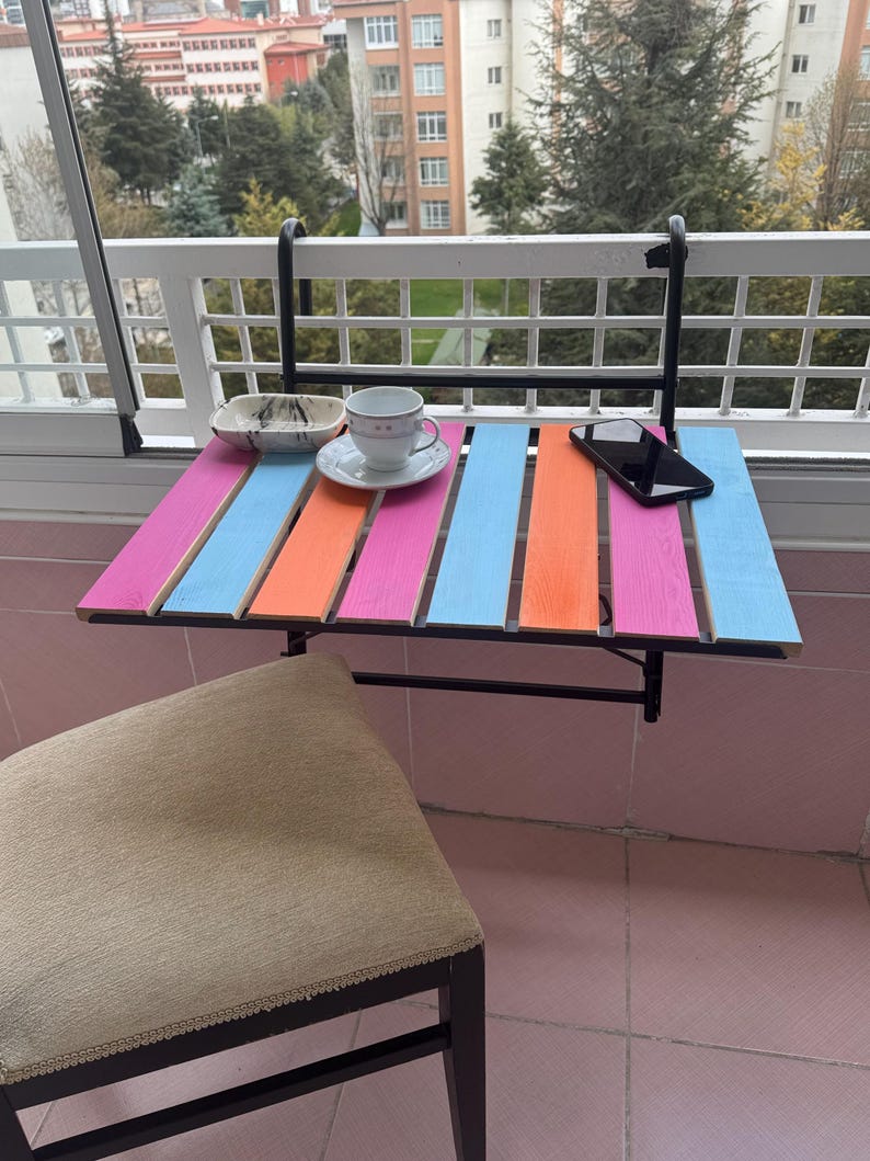 May include: A colorful folding table with a black metal frame and a pink, orange, and blue striped top. The table is attached to a white railing and has a small white cup and saucer on it. A black cell phone is also on the table.