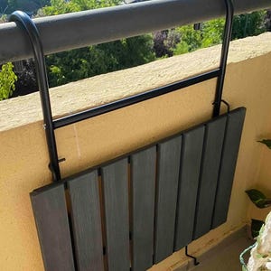 Puede incluir: Una mesa plegable de metal negro con una tapa de madera gris oscuro, montada en una barandilla de balc&oacute;n. La mesa est&aacute; plegada y asegurada con ganchos.
