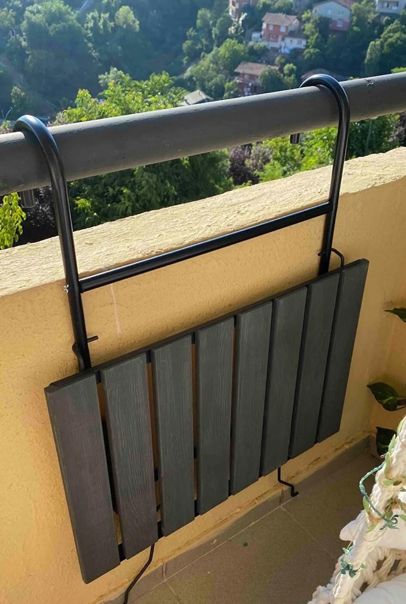 Puede incluir: Una mesa plegable de metal negro con una tapa de madera gris oscuro, montada en una barandilla de balc&oacute;n. La mesa est&aacute; plegada y asegurada con ganchos.