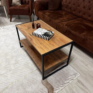 Könnte beinhalten: Ein rechteckiger Couchtisch mit einer hellbraunen Holzplatte und einem unteren Regal, umrahmt von einer schwarzen Metallstruktur. Ein Buch mit schwarz-weiß gestreiften Kanten, eine Kerze und eine kleine Flasche liegen auf dem Tisch. Der Tisch steht vor einem braunen Sofa.