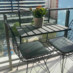 Puede incluir: Una mesa de patio de metal negro con dos sillas. La mesa tiene una tapa de madera gris y las sillas tienen cojines negros. Una planta en maceta con flores naranjas est&aacute; sobre la mesa.