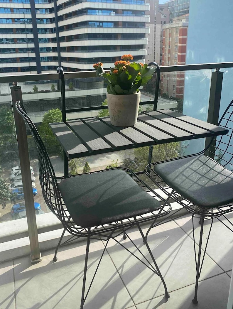 Puede incluir: Una mesa de patio de metal negro con dos sillas. La mesa tiene una tapa de madera gris y las sillas tienen cojines negros. Una planta en maceta con flores naranjas est&aacute; sobre la mesa.