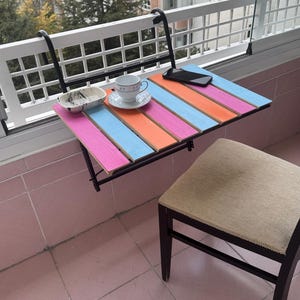 May include: A colorful folding table with a black metal frame attached to a balcony railing. The table has pink, blue, and orange stripes. A white cup and saucer and a cell phone are on the table.