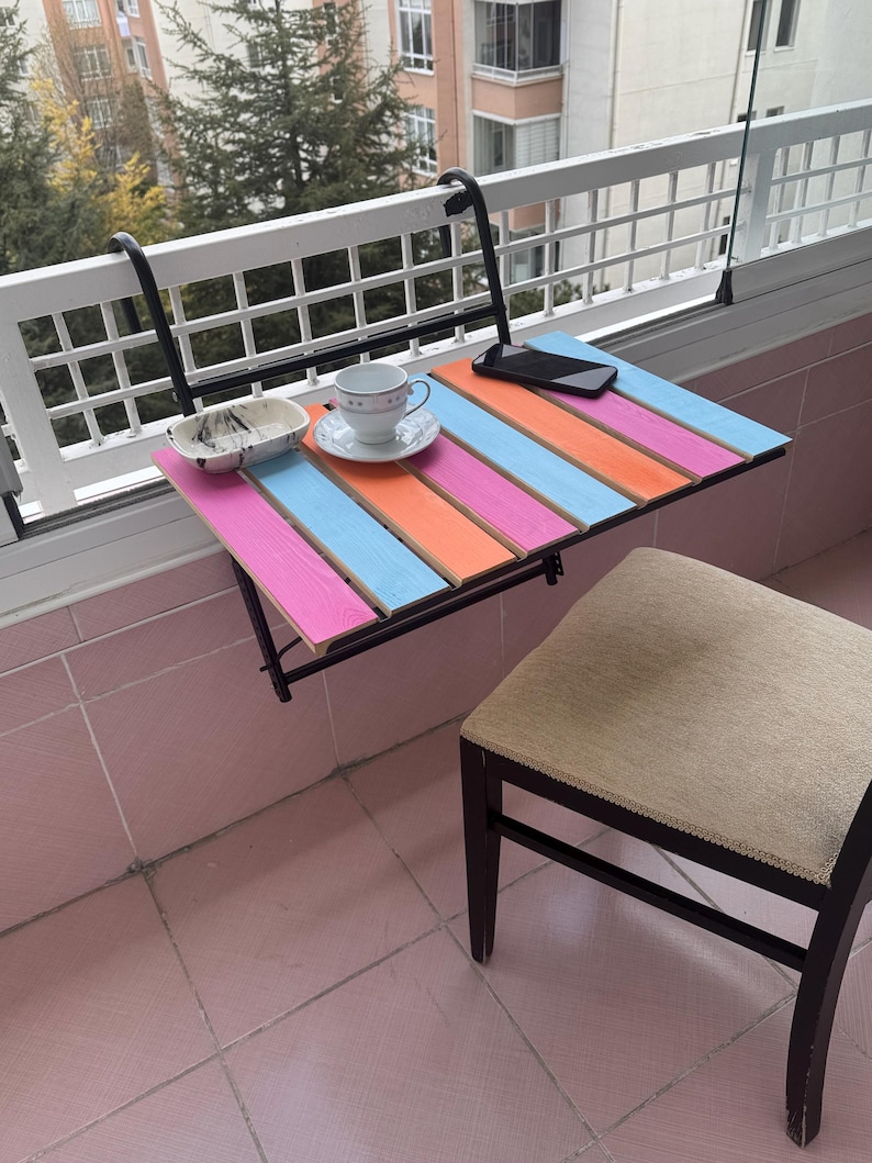 May include: A colorful folding table with a black metal frame attached to a balcony railing. The table has pink, blue, and orange stripes. A white cup and saucer and a cell phone are on the table.