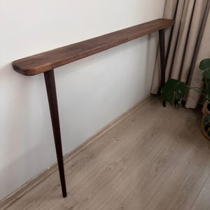 May include: A long, narrow console table constructed from dark brown wood. The table features a slim, rectangular tabletop with rounded edges and is supported by two slender, tapered legs that extend outward toward the floor.