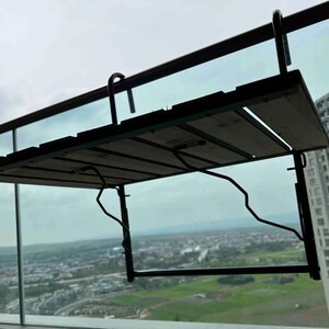 Puede incluir: Una mesa de balc&oacute;n de metal negro con una tapa de madera. La mesa est&aacute; plegada y unida a una barandilla. La mesa tiene vistas a una ciudad.