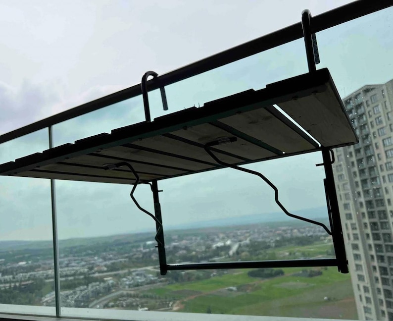 Puede incluir: Una mesa de balc&oacute;n de metal negro con una tapa de madera. La mesa est&aacute; plegada y unida a una barandilla. La mesa tiene vistas a una ciudad.