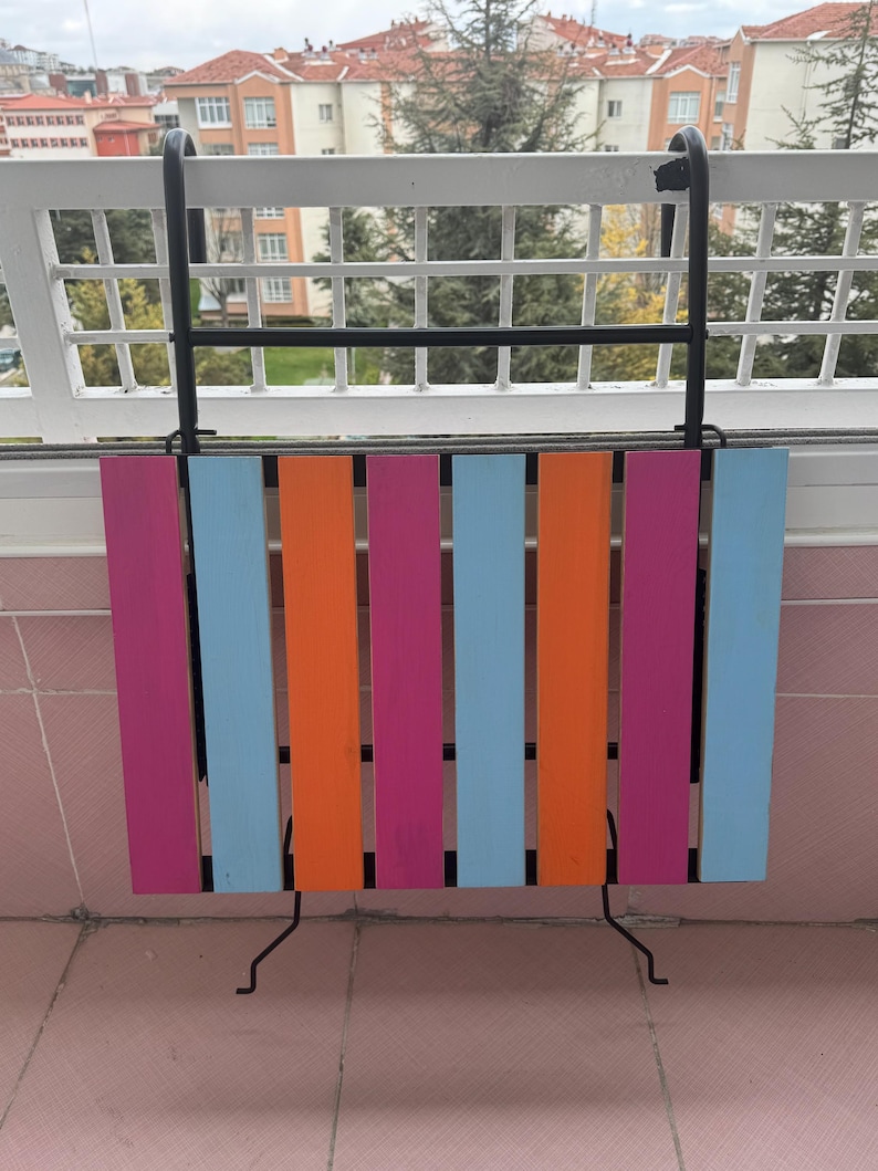 May include: A colorful folding table with a black metal frame. The table is made of wood and has alternating stripes of pink, orange, and blue.