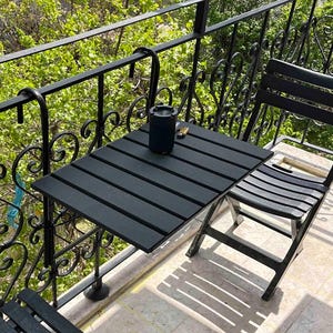 May include: Black metal balcony railing with a black folding table and two black chairs. The table is attached to the railing with brackets.