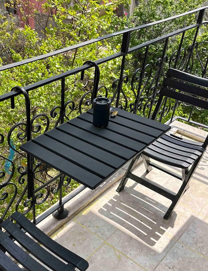 May include: Black metal balcony railing with a black folding table and two black chairs. The table is attached to the railing with brackets.