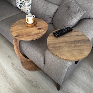 May include: Two round wooden side tables with a gray couch in the background. The table on the left has a white mug with a yellow sunflower design on it. The table on the right has a black remote control on it.
