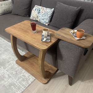 May include: A wooden side table with a curved leg design. The table has a rectangular top and a lower shelf. A small tray table is attached to the sofa. A glass of red liquid and a cup of coffee sit on the tables.