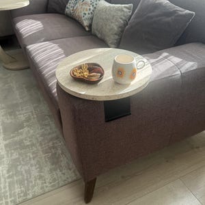 May include: A living room with a mauve sofa and grey cushions. A round side table with a marble top is attached to the sofa arm, holding a mug with a sunflower design and a wooden bowl with snacks. The sofa has wooden legs and a patterned throw pillow.