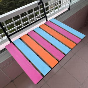 May include: A colorful folding table with pink, orange, and blue wooden slats. The table is attached to a black metal bracket and is mounted to a white railing.