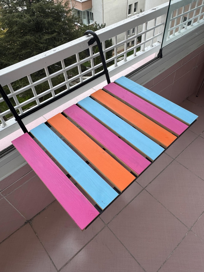 May include: A colorful folding table with pink, orange, and blue wooden slats. The table is attached to a black metal bracket and is mounted to a white railing.