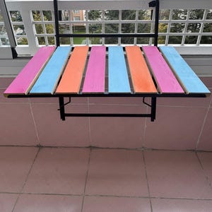 May include: A colorful folding table with a black metal frame. The table top is made of wood painted in alternating stripes of pink, blue, and orange.