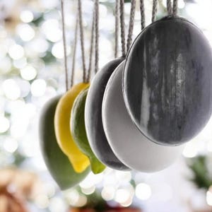 May include: A series of oval-shaped soaps hanging from individual jute strings. The soaps are arranged in a row and feature a variety of solid colours including dark grey, white, light grey, green, yellow, and olive green with smooth surfaces.