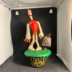 May include: A handmade clay figurine of a character from the video game "The Neverhood". The character is standing on a green base with the game's title written in gold. The character is wearing a red shirt and has a long, thin neck.