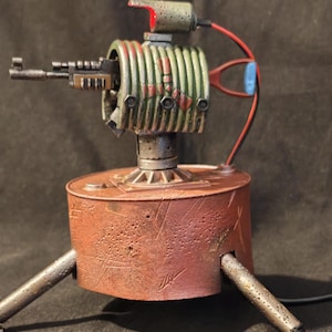 May include: A handmade, steampunk-style metal sculpture resembling a small turret gun. The sculpture features a rusty red base with three legs, a green and red gun barrel, and a red light on top. The piece has a detailed, industrial aesthetic.