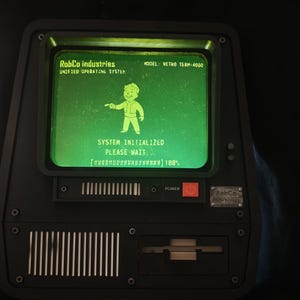Op de afbeelding: Een zwarte computerterminal met een groen scherm waarop RobCo Industries Unified Operating System, Model: Retro Term-4000, een Vault Boy-afbeelding en de tekst System Initialized Please Wait met een laadbalk op 100 procent staan.