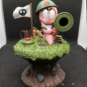 May include: A whimsical figurine of a pink worm soldier wearing a green helmet with a red star, holding a cannon. A white flag with a skull and crossbones stands nearby. The base is a green, grassy mound with a red button and a brown, tree-like structure.