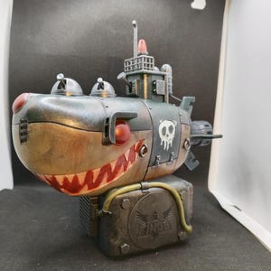 May include: A green, brown, and red submarine with a skull and crossbones on the side. The submarine is on a black base with a metal plate that says "KINGD" with a winged eagle logo.