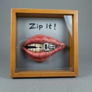 May include: A framed artwork featuring a sculpted mouth with a zipper across the lips. The lips are painted red with visible teeth, and the zipper is silver. The background is gray, and the text "Zip It!" is above the mouth. The frame is made of wood.