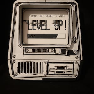 Può includere: Un cartello decorativo bianco e nero a forma di monitor di computer vintage. Lo schermo mostra il testo I DON'T GET OLDER, I JUST LEVEL UP! con una barra dell'esperienza etichettata come EXP. Il design presenta contorni neri disegnati.
