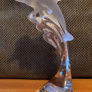 May include: A clear glass dolphin sculpture with a frosted finish, leaping out of a wave. The base of the sculpture is marked with the word "WACHTMANN".