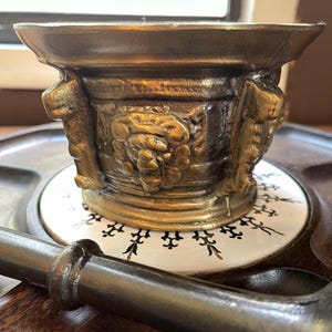 Op de afbeelding: Een vintage messing vijzel en stamper set. De vijzel heeft een brede rand en is versierd met reliëfs. De stamper is donker en glad. De set wordt gepresenteerd op een decoratieve keramische tegel.