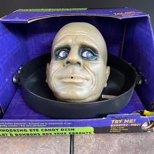 May include: A Halloween candy dish with a pale, bald head and blue eyes, set in a black dish. The dish is inside a purple box with the text "Wandering Eye Candy Dish". The box also has the text "Try Me!"