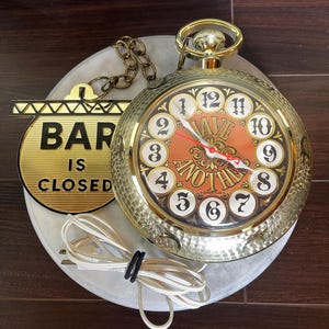 May include: A gold-toned wall clock shaped like a pocket watch with a textured metallic finish. The clock face features the text "HAVE ANOTHER" in red and black, with white numbers 1 through 12. A gold sign attached by a chain reads "BAR IS CLOSED".
