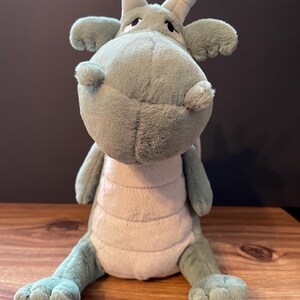May include: A soft toy dragon in green and beige. The dragon has large eyes, a soft snout, and two horns. The toy is sitting on a wooden surface.