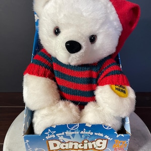 May include: A white plush bear wearing a red Santa hat and a red and dark green striped jumper. The bear is inside a blue cardboard box labelled Dancing Christmas Bear, which features the text Sing and Dance Along with Me and Jingle Bell Rock.