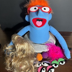 Könnte beinhalten: Eine blaue Handpuppe mit orangefarbenen Haaren und einer rot umrandeten Brille, mit offenem Mund und rosa Zunge. Das Zubehör umfasst eine blonde Perücke, eine rosa Federboa, eine orangefarbene Nase, eine violette Nase und mehrere Filzaugen.